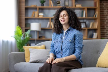 Kıvırcık saçlı bir kadın iç huzurunu bulurken kapalı gözlerle meditasyon yapıyor ve rahat bir ev ortamında rahat bir kanepede dinleniyor.