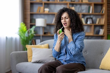 Evdeki kanepede oturan kadın boğaz spreyiyle boğaz ağrısı çekiyor. Soğuk algınlığından ya da grip belirtilerinden korunmak isterken rahatsız ve rahatsız görünüyor.