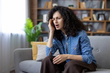 Kıvırcık saçlı genç bir kadın evdeki bir kanepede oturuyor, migrenden dolayı başını tutuyor, yorgun ve stresli, dinlenmek ve rahatlamak isteyen bir ev ortamında.