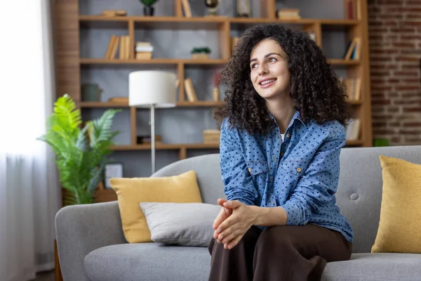 Kıvırcık saçlı, gülümseyen ve başka tarafa bakan bir kadın, arka planda bir kitaplıkla oturma odasındaki gri bir kanepede rahatça dinlenirken hayal kuruyor.