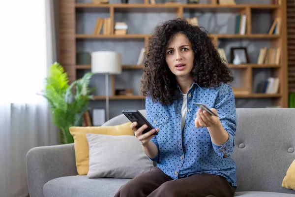 Evdeki kanepede oturan bir kadın, elinde bir cep telefonu ve kredi kartı ile çevrimiçi ödeme hatası ya da alışveriş yaparken yaşanan dolandırıcılık yüzünden kafası karışmış ve şok olmuş görünüyor.