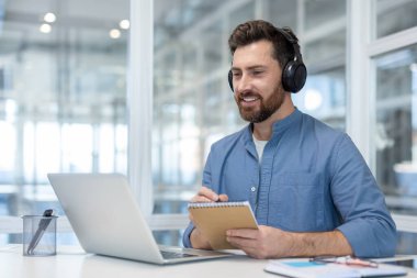 Kulaklık takan, gülümseyen sakallı bir adam bilgisayarında profesyonel bir web sitesi yayınlıyor, aktif olarak dinliyor ve bir ofis masasındaki bir not defterine notlar alıyor.