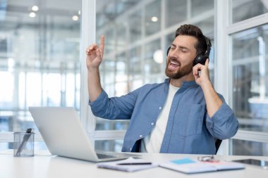 Mutlu sakallı bir adam, iş molasından zevk alıyor, kulaklık takıyor ve müzik eşliğinde şarkı söylüyor, modern ofis masasında dinlenirken, iş günlerinde neşe ve stres buluyor.