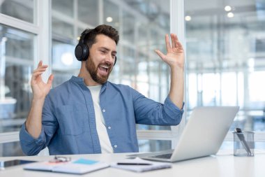 Kulaklık takan, müzikten zevk alan ve laptopunun başında çalışırken heyecanla şarkı söyleyen genç bir adam, başarıyı kutluyor ve kaygısız hissediyor.