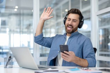 Kulaklık takan ve akıllı telefon tutan bir adam, ofis masasından gelen bir video konuşmasında elini sallayarak uzaktan çalışmayı ve sanal iletişimi sembolize ediyor.