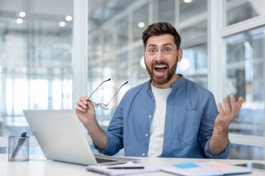 Modern ofis dizüstü bilgisayarındaki iş adamı sevinçli bir sürprizle karşılık veriyor ve terfi, anlaşma veya başarılı yatırım hakkında iyi haberler okuduktan sonra elini kaldırıyor.