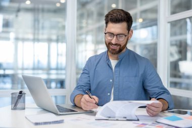 Masasında gülümseyen gözlüklü sakallı işadamı, modern bir ofiste dizüstü bilgisayarla çalışırken kontrat imzalıyor, profesyonel bir ortamda odaklanmış ve kendinden emin.