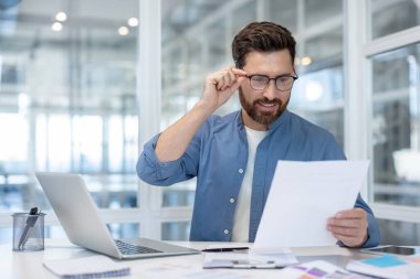 Genç işadamı, önemli bir yasal belge veya iş raporunu okurken gözlüğü ayarlıyor, bilgiyi analiz ediyor ve modern bir ofis ortamında çalışıyor