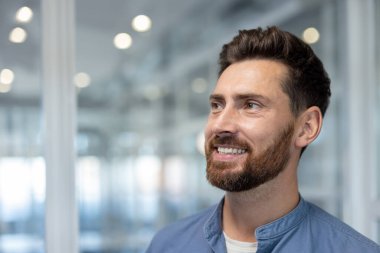 Mavi tişörtlü, beyaz tişörtlü, kendine güvenen sakallı bir adam, modern ofis ortamında düşünceli bir ifadeyle yüzünü kaçırırken gülümsüyor.