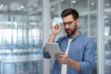 Modern bir ofiste duran genç adam, dijital tablet içeriğini dikkatle okurken gözlüklerini ayarlıyor, modern iş iletişimi ve teknoloji kullanımını yansıtıyor.