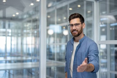 Gözlüklü gülümseyen işadamı elini uzatıyor, selamlıyor ve modern ofis ortamında yeni bir meslektaşını veya müşterisini selamlıyor.