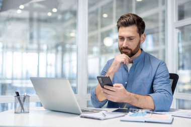 Modern bir ofiste çalışan bir iş adamı, akıllı telefonuna odaklanırken masada bir laptop açık, üretkenlik, teknoloji ve iletişim kavramlarını temsil ediyor.