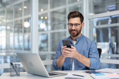 Gözlüklü ve sakallı gülümseyen bir işadamı, bir masada oturmuş, akıllı bir telefonla mesajlaşırken neşeli görünüyor, bir dizüstü bilgisayar ve çağdaş ofis masasının üzerindeki belgelerle.