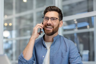 Modern bir ofiste çalışırken cep telefonuyla sohbet eden, güler yüzlü, sakallı bir işadamı kendinden emin, arkadaş canlısı ve profesyonel bir görüşmeyle meşgul görünüyor.