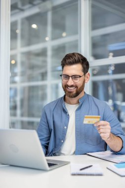 Gülümseyen adam çevrimiçi alışveriş yapmaktan zevk alıyor, bilgisayarını kullanıyor ve modern ofis masasında güvenli bir e-ticaret ödemesi yapmak için bir kredi kartı tutuyor.