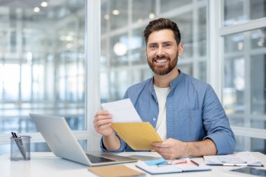 Kahverengi bir zarftan gelen önemli bir mektubu açan gülümseyen adam, beyaz bir ofis masasında dizüstü bilgisayar ve belgelerle oturuyor, olumlu haberler alıyor veya başarılı bir iletişim kuruyor.