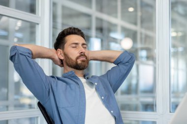 Sakallı ve gözleri kapalı genç bir adam sandalyeye yaslanıyor, modern bir ofiste dinleniyor, huzurlu bir stres atağı ve zihinsel sakinliğin tadını çıkarıyor.