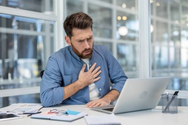 Modern ofisteki dizüstü bilgisayarın başındaki genç adam göğsünü tutuyor. Ani göğüs ağrısı ve nefes darlığı yüzünden yüzünü buruşturuyor. İş yerinde endişeli ve sıkıntılı görünüyor.