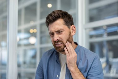 Sakallı bir adam keskin diş ağrısı çekiyor, yanağını eliyle tutuyor, kaşlarını çatıyor, diş probleminden ya da oral sağlıktan rahatsız olduğunu ifade ediyor.