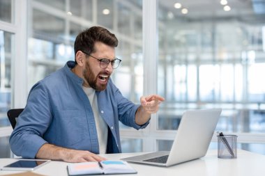 Kızgın işadamı, başarısız bir proje, teslim tarihi ya da teknik bir sorun yüzünden hayal kırıklığı ve stresi gösteren modern bir ofis masasına bağırıyor ve dizüstü bilgisayarına işaret ediyor.