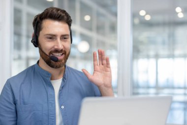 Mikrofonlu bir kulaklık takmış gülümseyen sakallı bir adam, bir video konferansına katılıyor ya da parlak, modern bir ofis ortamından giyiniyor.