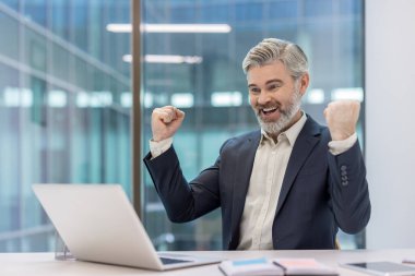 Olgun iş adamı zaferle yumruklarını kaldırıyor, finansal başarısını ve online başarısını kutlarken modern kurumsal ofisinde bir dizüstü bilgisayara bakıyor.