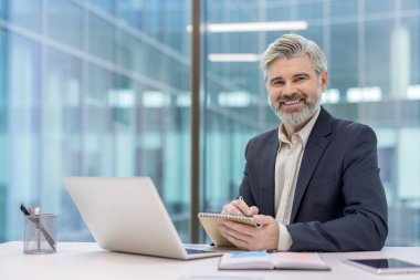 Ofis masasında oturan gri saçlı ve sakallı olgun bir iş adamı dizüstü bilgisayarın yanına notlar yazarken kameraya güvenle gülümsüyor.
