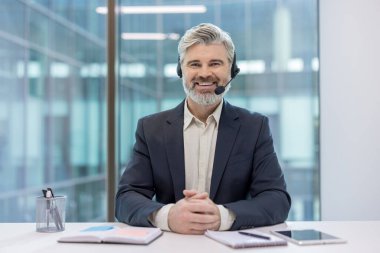 Kulaklık ve takım elbise giyen gülümseyen kıdemli bir adam, elleri bağlı bir ofis masasında oturuyor, müşteri hizmetlerini, teknik desteği ve çağrı merkezinin profesyonelliğini temsil ediyor.