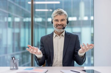 Modern bir ofisteki video görüşmesinde, müşteri desteği, sanal danışmanlık ya da tele pazarlama hizmetlerini temsil eden, kulaklık ve mikrofonla gülümseyen olgun iş adamı.