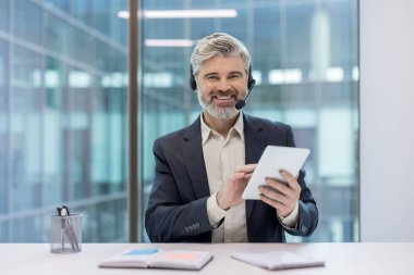 Modern bir ofiste çalışırken kulaklık takan gülümseyen iş adamı, çevrimiçi müşteri desteği sağlıyor ve dijital tablet kullanarak müşterilerle iletişim kuruyor.