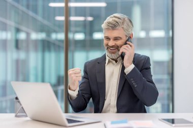 Yetişkin iş adamı akıllı telefondan iyi haberler alıyor, modern bir ofis masasında bilgisayarla çalışırken muzaffer bir yumruk hareketi yapıyor, neşe ve başarıyı ifade ediyor.