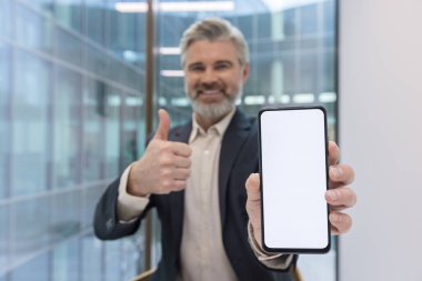 Üst düzey işadamı elinde boş beyaz ekran maketi olan bir akıllı telefon tutuyor, gülümsüyor ve baş parmak işareti yapıyor, onaylama veya tavsiye gösterisi yapıyor.