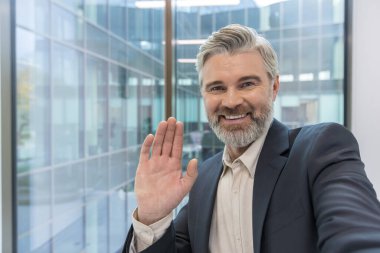 Modern bir ofisten sanal bir video konferansı sırasında kameraya gülümseyen ve el sallayan olgun bir yönetici, uzak bir toplantıda meslektaşlarıyla bağlantı kuran kendinden emin bir profesyonel.