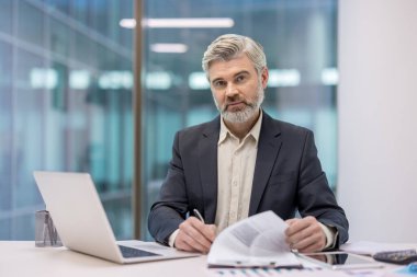 Ofis masasında çalışan olgun bir işadamı, belgeleri inceliyor ve bir dizüstü bilgisayar kullanıyor, profesyonellik, deneyim ve kurumsal sorumluluk kavramlarını temsil ediyor.