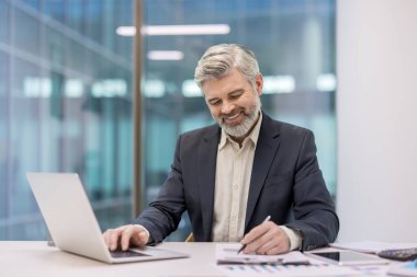 Ofis masasında çalışan kıdemli işadamı, dizüstü bilgisayarda yazı yazıyor ve belgeler üzerine notlar yazıyor, mali tabloları ve verileri incelerken gülümsüyor.