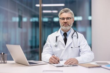 Beyaz laboratuvar önlüğü ve steteskop giyen olgun, gri saçlı bir doktor, bir masada dizüstü bilgisayarla oturuyor, kameraya bakıyor ve notlar alıyor, sağlık uzmanlığını somutlaştırıyor.