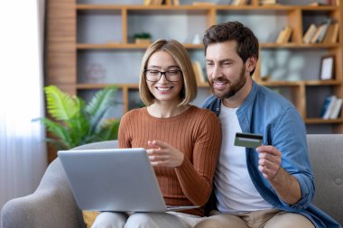 Genç mutlu çift internetten kredi kartıyla ödeme yapıyor, bir e-ticaret mağazasından eşyalar alıyor ve evdeki kanepede dinlenirken modern internet bankacılığının tadını çıkarıyor.