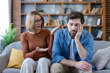 Çift koltukta tartışıyor, kadın hayal kırıklığını ifade ederken erkek üzgün ve bitkin görünüyor, aile ortamlarında ilişki sorunları ve iletişim sorunları gösteriyor.