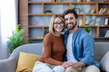 Mutlu genç çift gülümsüyor, modern oturma odalarında rahat bir koltukta kucaklaşıyorlar, yakın bir anın tadını çıkarıyorlar ve doğrudan kameraya bakıyorlar.