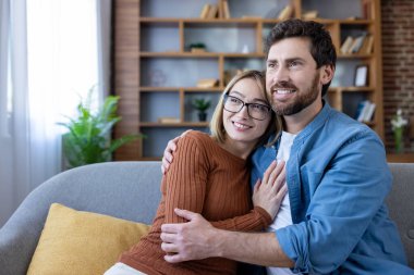 Sevgi dolu bir çift, rahat bir koltukta oturuyor, sarılıyor ve gülümsüyor, mutlu ifadelerle gözlerini kaçırıyor, konforu somutlaştırıyor, güçlü bir ilişki kuruyor ve modern bir oturma odasında ortak bir gelecek paylaşıyor.
