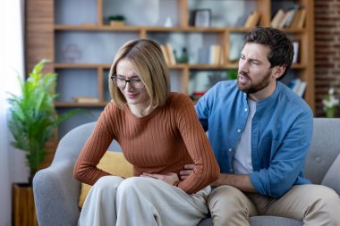 Kanepede otururken karın ağrısı ya da kramplar hisseden bir kadın. Endişeli bir adam onu rahatlatıyor ve oturma odasında destek öneriyor.