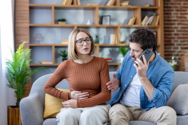 Kanepede şiddetli karın ağrısı çeken, karnını tutan bir kadın. Endişeli bir adam telefon eder. Muhtemelen durumu için tıbbi yardım ister.