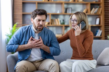 Göğüs ağrısı çeken ve göğsünü sıkıca tutan bir adam, endişeli bir kadın kanepesinde oturmuş, acil tıbbi yardım için akıllı telefonundan acil çağrı yapıyor.