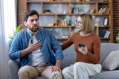 Bir çift evdeki kanepede oturuyor, erkek göğüs ağrısı çekiyor ve nefes almakta zorlanıyor. Kadın elinde akıllı telefon tutuyor, şok edici, stresli bilgilere ya da sağlık sorunlarına tepki veriyor.