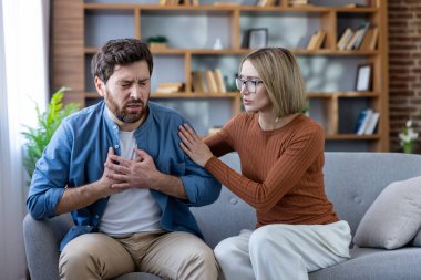 Akut göğüs ağrısı çeken ve bir koltukta otururken göğsünü iki eliyle tutan bir adam, elini omzuna koyarak onu destekleyen endişeli bir kadın.