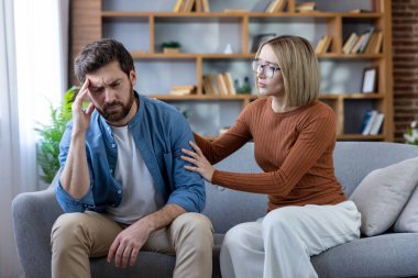 Stres ve baş ağrısı çeken bir erkeği destekleyen ve rahatlatan bir kadın, ev ortamında bir kanepede oturan, empati ve ilişki zorluklarını sembolize eden bir kadın.