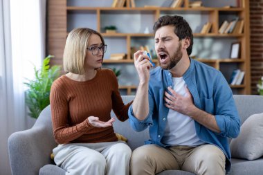 Astım krizi geçiren ve nefes almak için mücadele eden erkek, endişeli bir kadın destek sunarken, sağlık sorunlarını ve solunum sıkıntısını vurgularken astım spreyi kullanıyor.