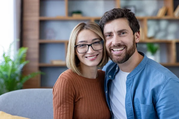 Genç bir adam ve kadın sıcakkanlılıkla kucaklaşıyor, mutlu bir gülümsemeyle kameraya bakıyor, evdeki bir kanepede dinlenirken, mutluluğu, ilişkileri ve ortaklığı temsil ediyor.