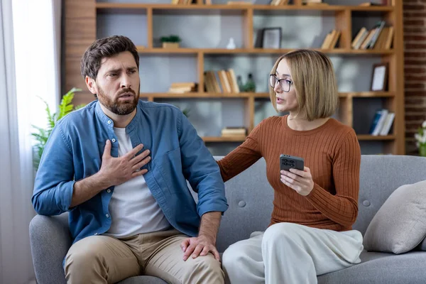 Bir çift evdeki kanepede oturuyor, erkek göğüs ağrısı çekiyor ve nefes almakta zorlanıyor. Kadın elinde akıllı telefon tutuyor, şok edici, stresli bilgilere ya da sağlık sorunlarına tepki veriyor.