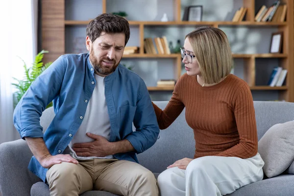 Adam karnını görünür bir acı içinde kanepeye bastırırken endişeli karısı yanında oturuyor ve bir evde oturma odasında ona destek oluyor.